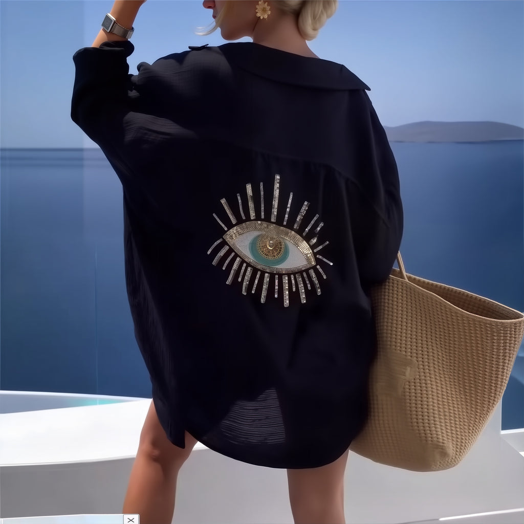 Black oversized shirt with embroidered eye motif worn over bikini by ocean, Scandinavian swimwear cover-up