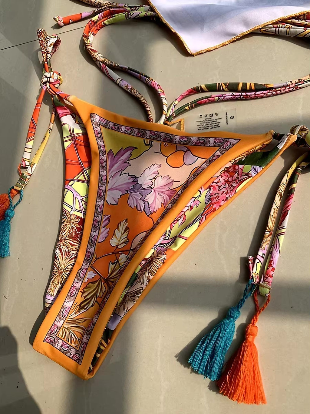 Orange floral bikini top with purple and multicolor botanical print, matching adjustable straps and tassel details.