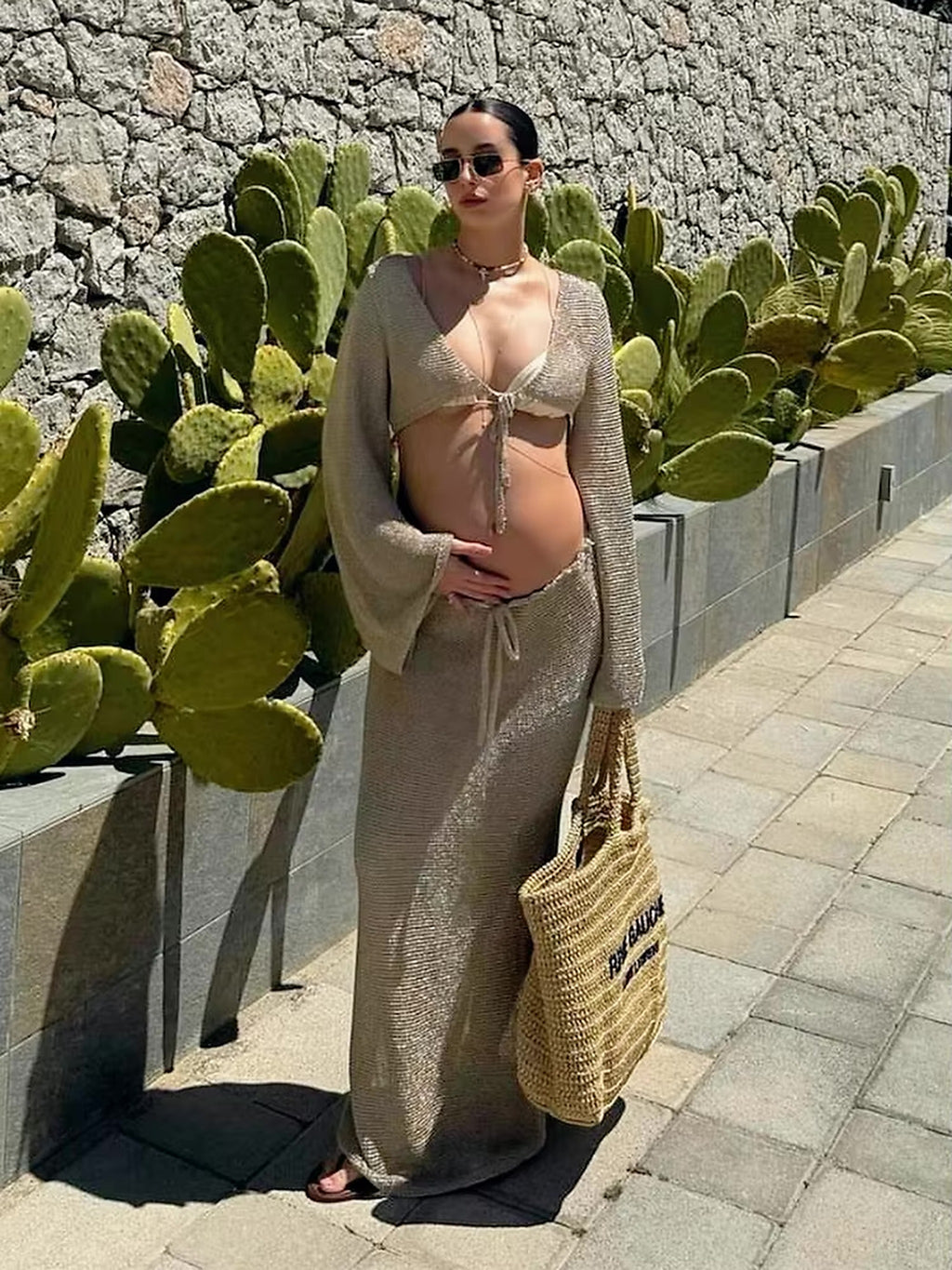 Pregnant woman wearing beige knit bikini top with tie-front detail and matching cover-up, posing by cacti against stone wall.