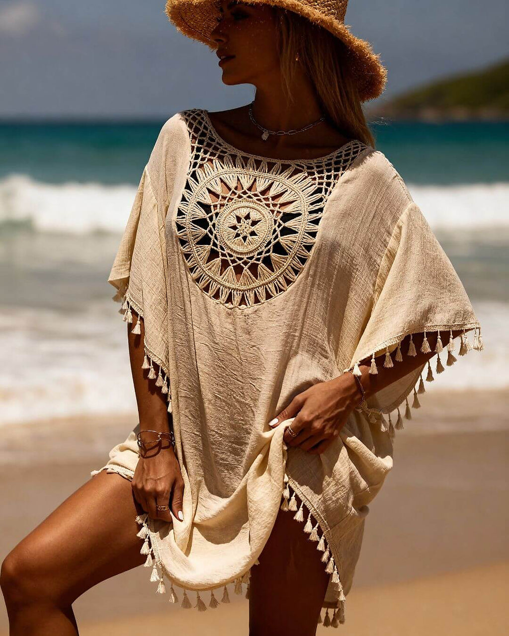 Woman wearing cream crochet beach cover-up dress with tassel trim and mandala pattern, standing on sandy beach with ocean
