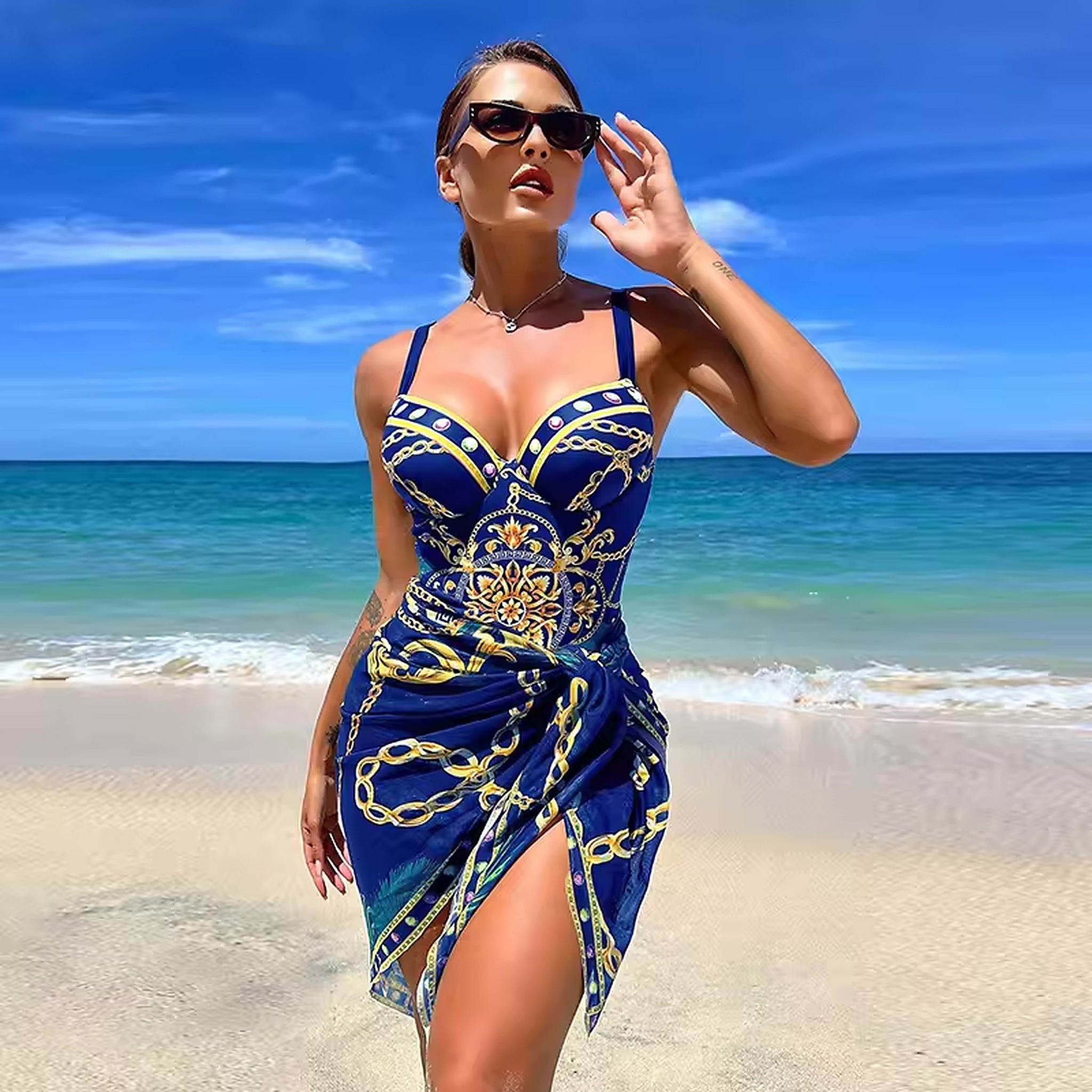 Woman wearing blue and gold chain print bikini with sarong wrap on beach with ocean and clear sky