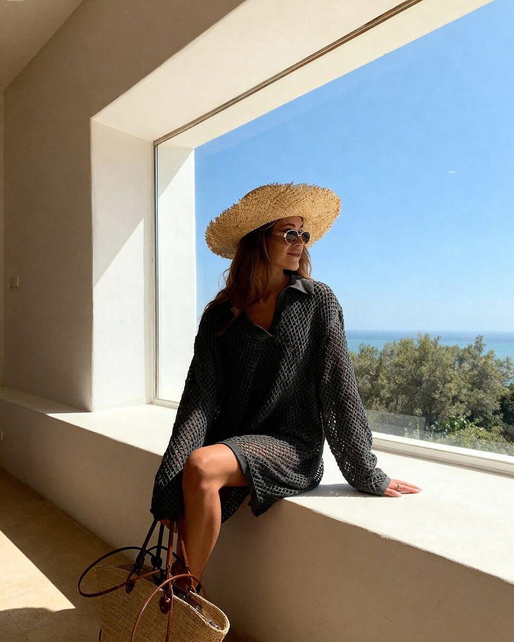 Woman wearing black textured bikini with knit cover-up, straw hat, and sunglasses by sunny window overlooking ocean