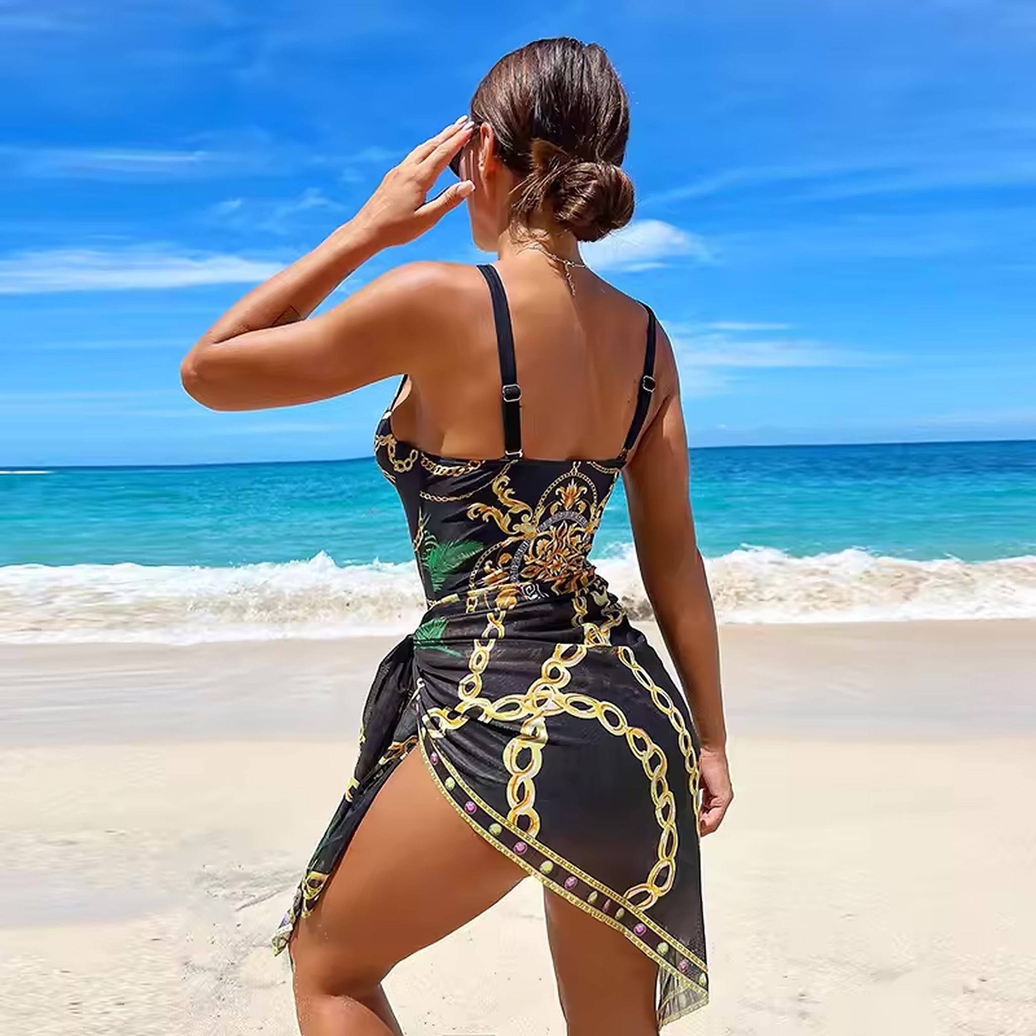 Woman wearing black bikini with gold chain print design on beach, ocean and blue sky in background