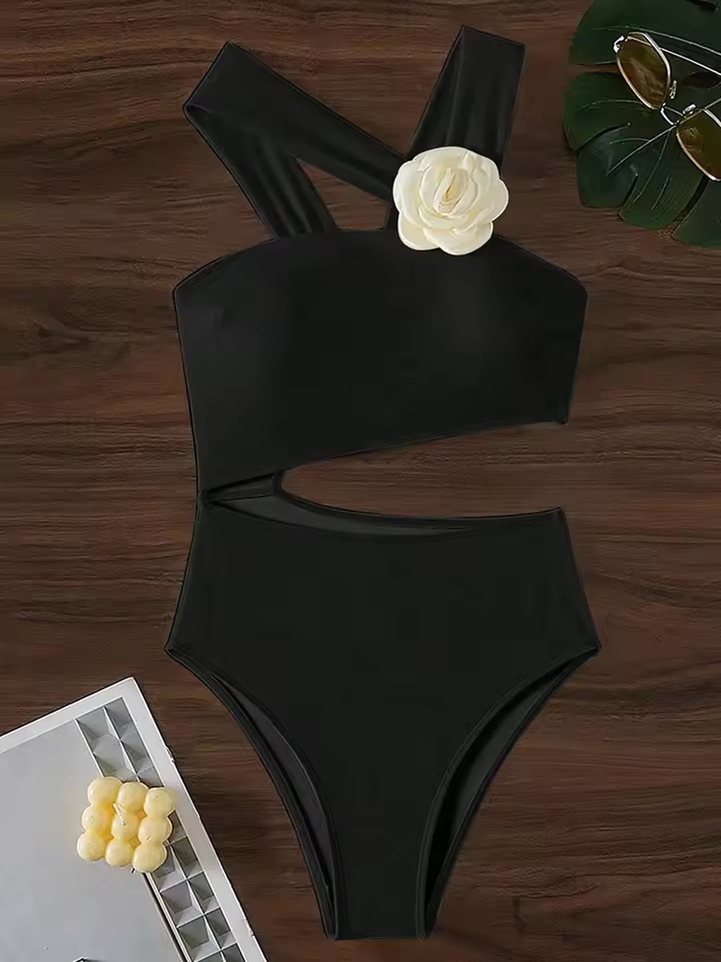 Black cutout one-piece swimsuit with shoulder straps, displayed flat lay on wooden surface with white flower accent